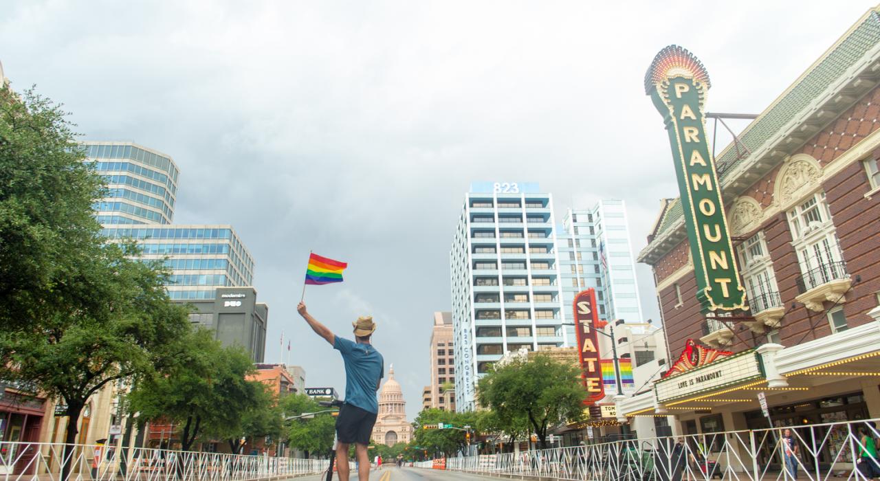 Man in faux cowboy hat waves rainbow flag on scooter in front of the Paramount Theater and Capitol Building